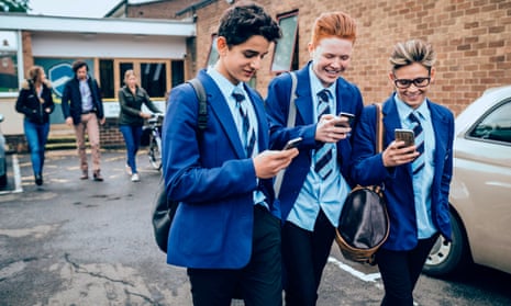Three schoolboys leaving school with their teachers behind them. They are all holding mobile phones and smiling; they wear bright blue blazers, pale blue shirts and blue ties