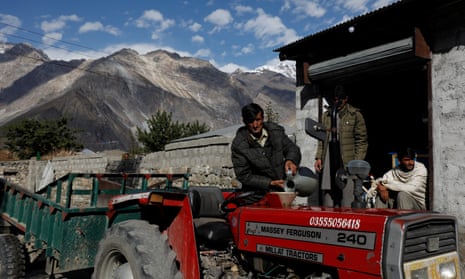 A man fills a bright red tractor with petrol in a mountain village, with steep mountain slopes behind him, framed against blue sky with little fluffy white clouds