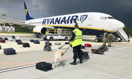 A guard in a hi-vis jacket holds a sniffer dog on a lead, checking bags laid out on the runway in front of a Ryanair plane
