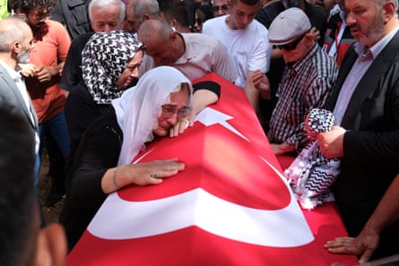 A woman cries over a coffin draped in a red and white Turkish flag while surrounded by people.