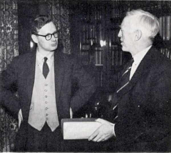 Whatmore standing with hands on hips looking at Monkhouse, who is holding a large book and talking to him, both wearing suits
