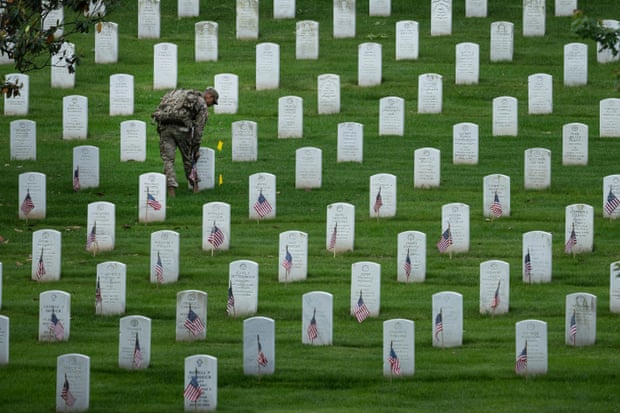 A man in military fatigues stands among rows of small white headstones placing small American flags.