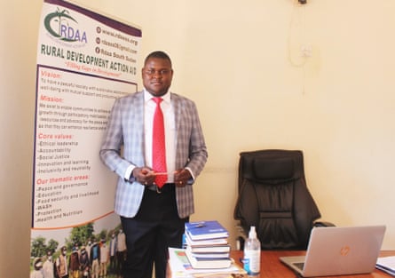 A man in a suit stands next to his desk.