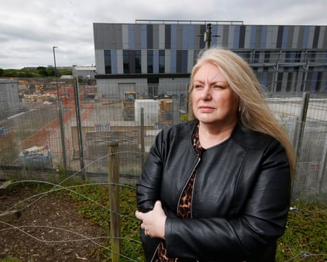 Diane Davidson in a black leather jacket stands by a wire fence with a construction site and modern building behind her