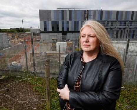 Diane Davidson in a black leather jacket stands by a wire fence with a construction site and modern building behind her