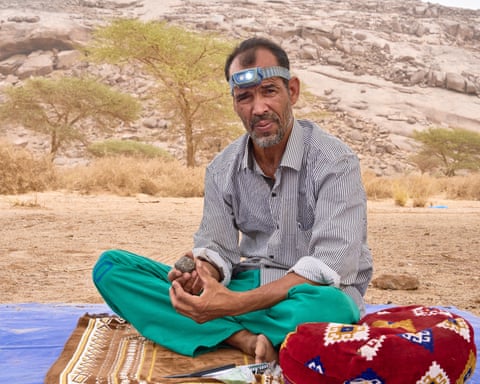 Lamine Ould Hanoun, sat on a carpet outdoors with a torch on his head holding a rock he is inspecting