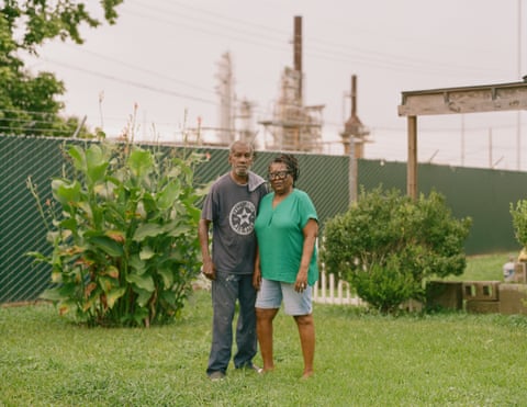 A middle-aged man and woman stand in a neatly groomed yard while an oil refinery looms in the background.