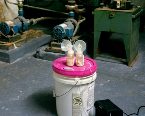 A pair of breast milk pumps half full of milk sit on top of an industrial sized chemical bucket, in an industrial looking small space.
