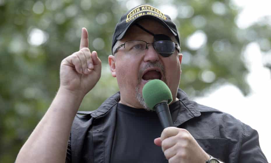 A man wearing an Oath Keepers hat and a plastic eye patch speaks into a microphone.