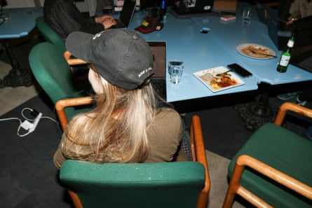 Someone with long blond hair wearing a black baseball cap is seen from behind sitting in a green armchair with wooden armrests, in front of a blue table with plates of food and bottles.