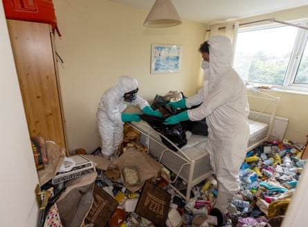 Two people wearing hazmat gear cleaning a room full of rubbish.