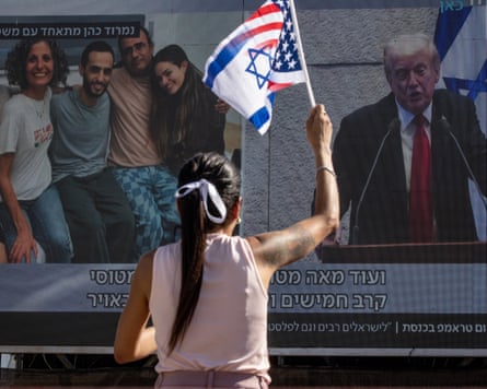 Mulher agita uma bandeira que é uma mistura das bandeiras de Israel e dos EUA enquanto olha para uma tela grande na rua mostrando a libertação dos reféns e Donald Trump falando