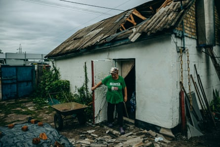 A middle-aged woman with a headscarf and wearing a green T-shirt with ‘volunteer’ printed on it is standing in a doorway of a house surrounded by debris and a whole in the roof