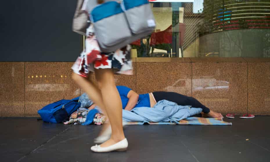 A homeless person is sleeping on the street during the day and a pedestrian is walking in front of him/her.