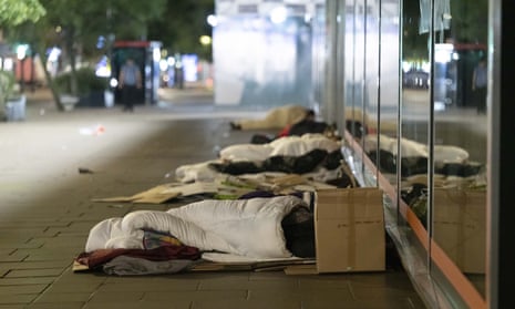 About five people sleep in a row outside central London shops at night. They have basic bedding including coverless blankets and a cardboard box for belongings