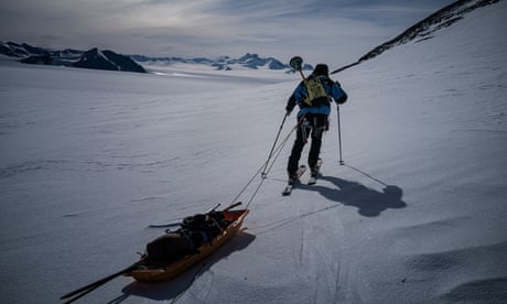 A man stands on a rocky outcrop looking out at a large snowy plain with snowy mountain peaks in the background