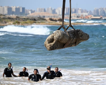 Divers in the sea watch as a statue is hoisted by a crane from the water