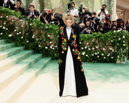 Anna Wintour in a black floral gown with a bank of photographers behind her.