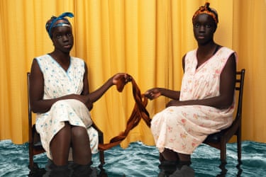 Two women in headscarves sit on chairs in water, connecting braided hair between them