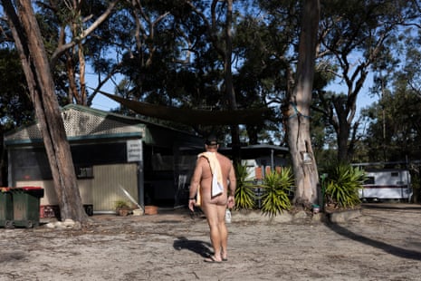 Naked Mick walks across a clearing with a towel over his shoulder.