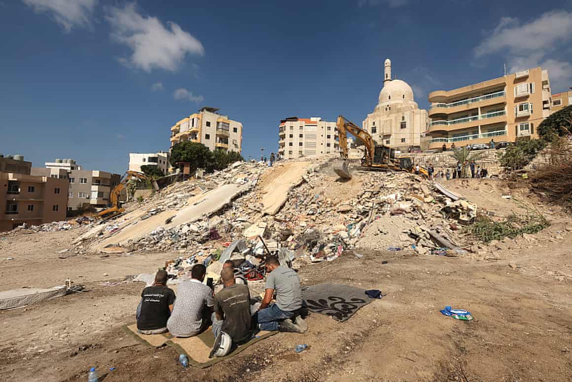 Men sit on rugs close to an area destroyed by Israeli airstrike