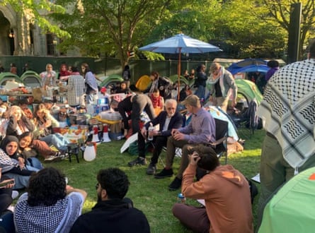 two men seated, talking, in the center, surrounded by students seated on the grass