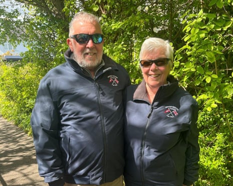 Alan and Jane Kelvey in matching sailing jackets stand together outdoors in front of leafy trees