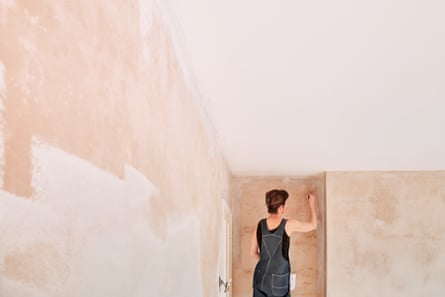 Woman in overalls painting a newly plastered wall with a roller brush.
