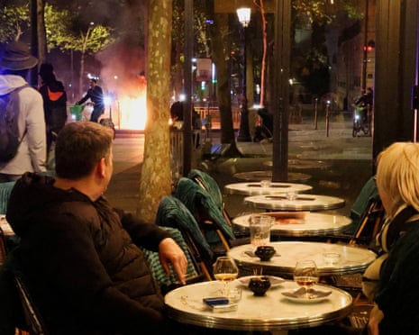 Paris cafe patrons sit at outdoor tables watching flames from a riot fire nearby