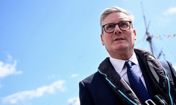 Keir Starmer visits Portsmouth: he is seen in a head and shoulders photo standing on the deck of a historic ship, with its mast in the background. He is looking slightly upwards and is framed against the blue sky; he wears a dark suit and tie, white shirt and a life-vest.
