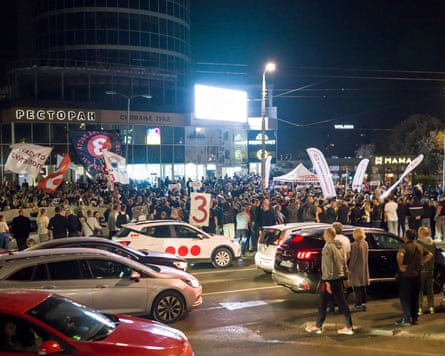 A crowd gathers at a nighttime intersection with flags and parked cars