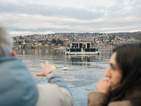 Two out-of-focus people in the foreground look towards a lake with a floating platform and building on it.
