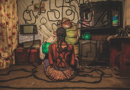 A seated woman, seen from behind in a living room, with braided hair extensions running like cables from her head over the floor and walls