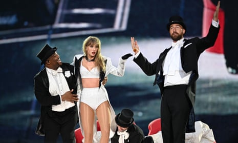 Two men wearing black suits and top hats stand on either side of Swift, wearing a white jacket and two-piece set on stage