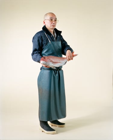 A bald man in glasses and an apron holds a large red fish with both hands