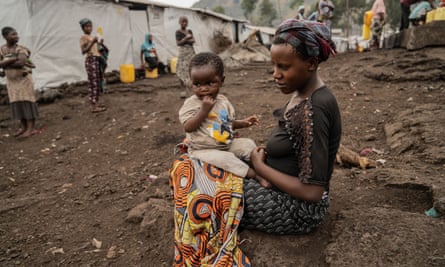 A baby sits on her mother’s knee in a displacement camp. She has scars from mpox and has a blanket around her knees