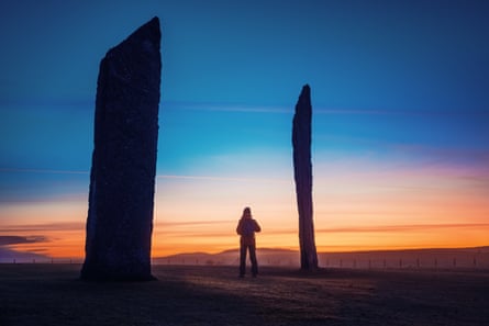 A figure standing between two tall standing stones with a dramatic sunset behind