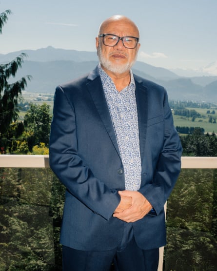 Avtar Singh standing with his hands clasped in front of him, wearing a blue suit and a blue-and-white patterned shirt, against a background of greenery with mountains in the distance