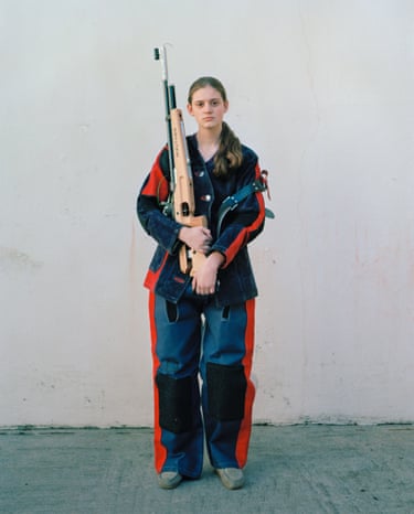 Teenager in heavy sports gear cradles a large rifle staring into camera