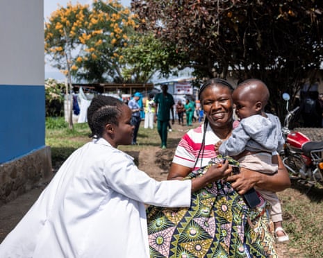 A smiling woman holds a baby boy in her arms while a nurse looks on and a group of people gather behind her.