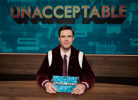 Ed Gamble sits at a desk holding a card, smiling, with the Unacceptable show logo behind him