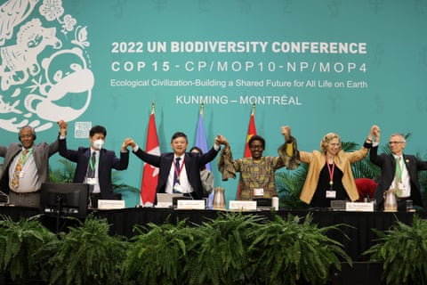 Six people stand holding hands in front of a sign saying "2022 UN Biodiversity Conference" qeituidqriqrhinv