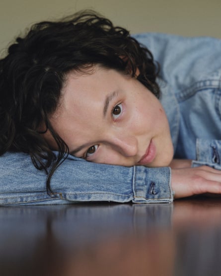 A closeup of Bella Ramsey wearing a denim jacket and leaning her head on her bent right arm, on a shiny brown table