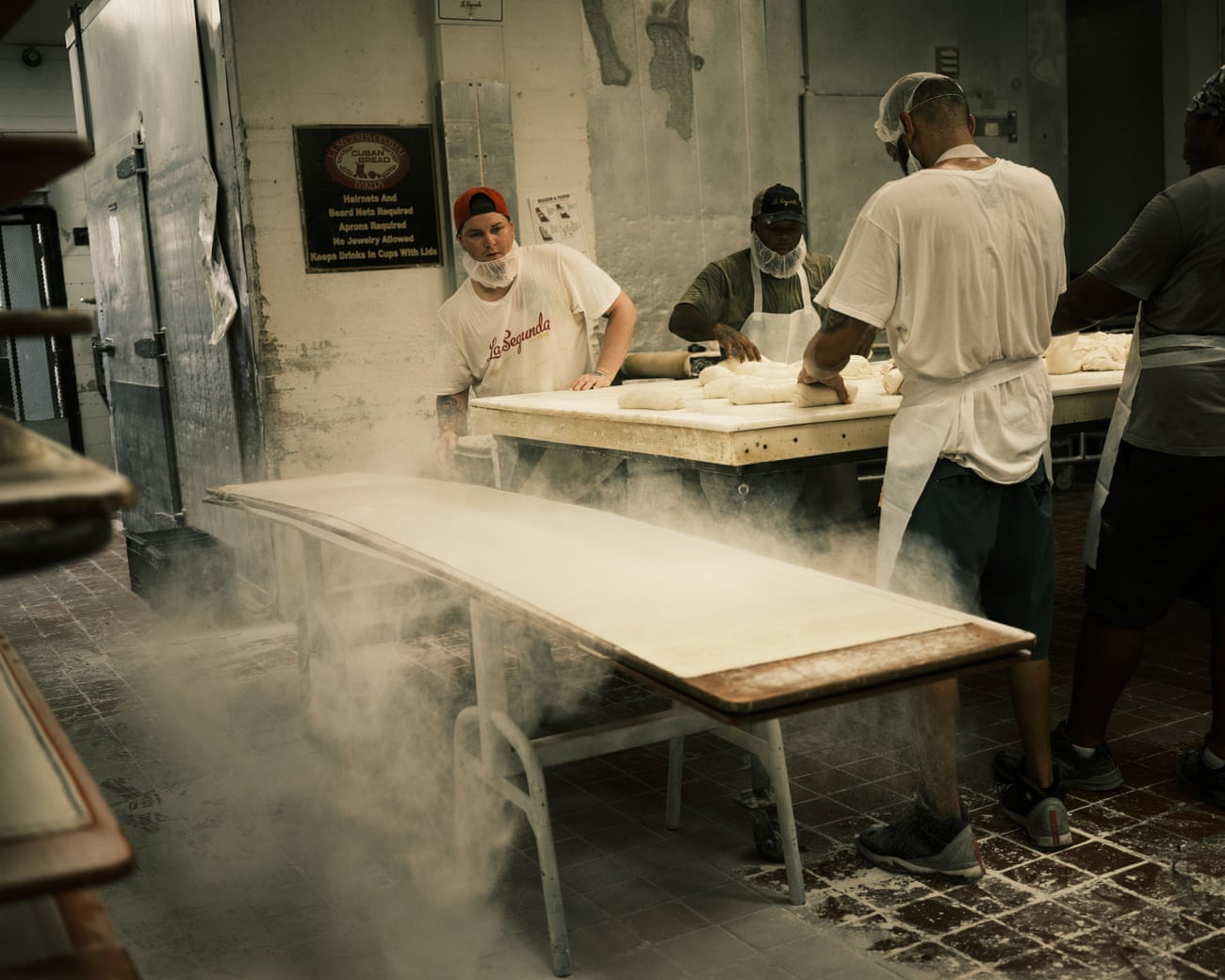 A man throws flour onto a long wooden table while two others stand at another table handling dough in the kitchen of a commercial bakery.