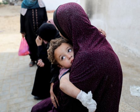 A woman wearing a traditional headscarf holds a child with a bandaged hand as others bow their heads in mourning in the background