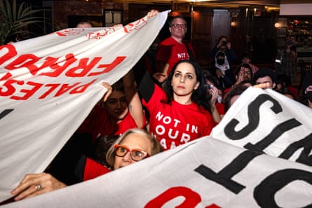 people hold banners while wearing red shirts that say ‘not in our name’