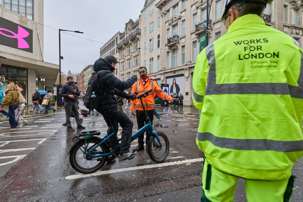 A young man in a black hooded anorak, on a blue folding electric bicycle, is halted in the street by a man in an orange hi-vis jacket.