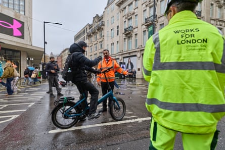 A young man in a black hooded anorak, on a blue folding electric bicycle, is halted in the street by a man in an orange hi-vis jacket.