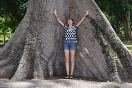 Bonnie Waring stands with her hands in the air leaning against the bottom of a gigantic tree.