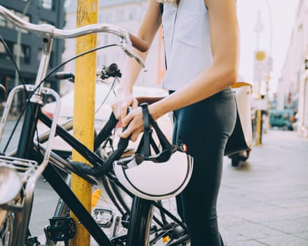 A person locking bicycle with pole on sidewalk in city. Their cycle helmet is dangling from their wrist. It appears to be a bright sunny day.